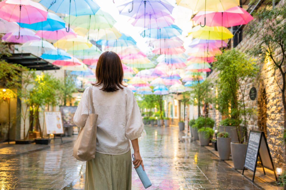 雨上がりの街で折りたたみ傘を手に歩く人の頭上を、色とりどりのアンブレラスカイが彩る風景