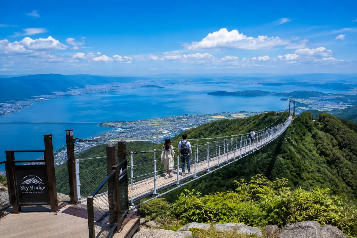 青空と琵琶湖を一望しながら山上のSky Bridgeを渡る旅行者の様子