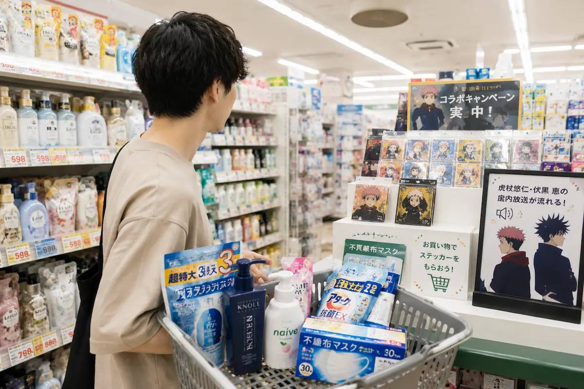 日用品を入れた買い物かごを持ちアニメコラボ売り場を見る男性